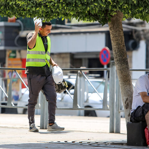 A man browses a phone in the shade of a tree while a construction worker shields his head from the sun with a water bottle while walking along a street in Algiers on July 18, 2023 during a heat wave. (Photo by AFP) (Photo by -/AFP via Getty Images)